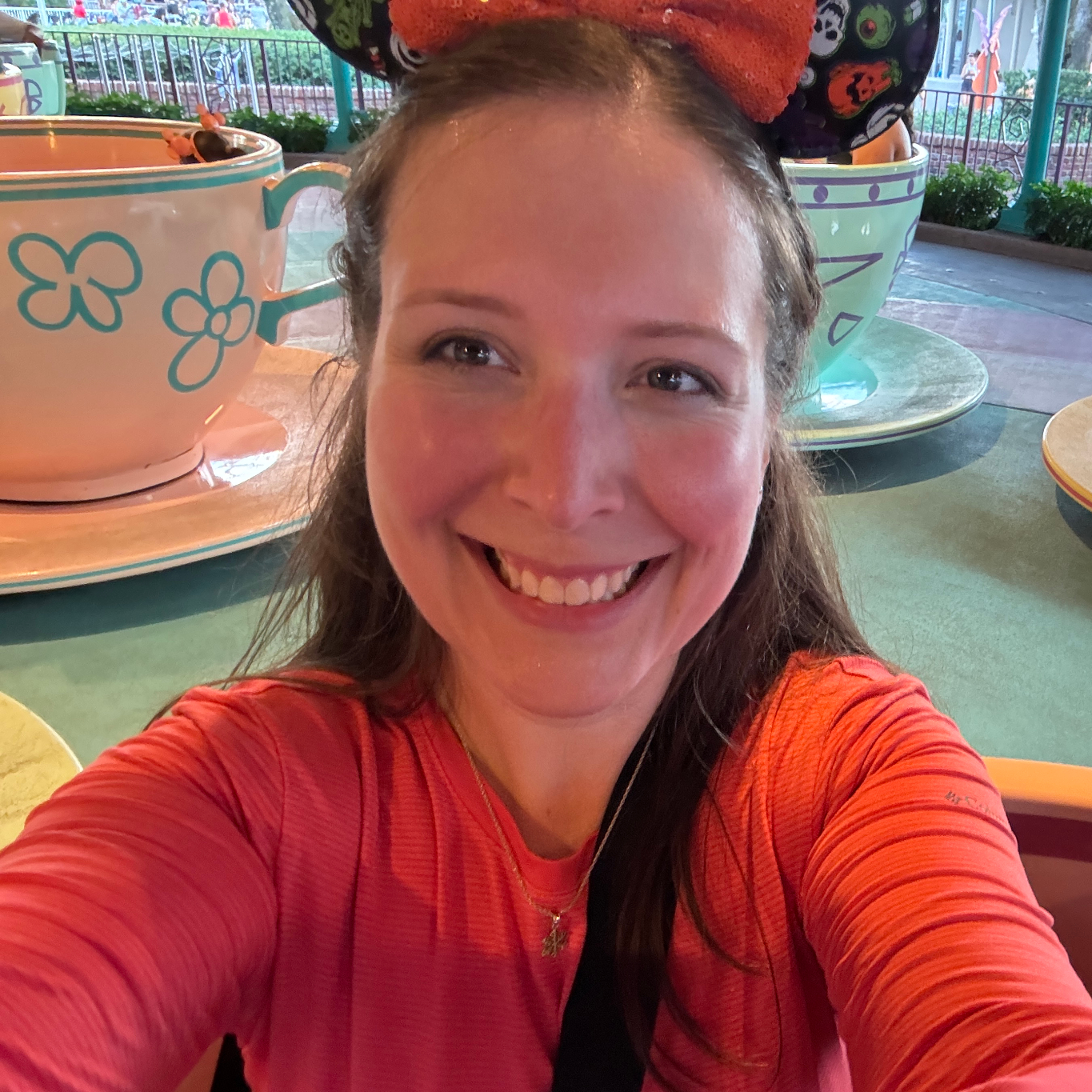 Kristy Lacroix Disneyworld 2024. Kristy is smiling brightly while taking a selfie on the Mad Tea Party ride at Walt Disney World. She is wearing a red-orange long-sleeve shirt and sparkly orange Minnie Mouse ears with a Halloween-theme.